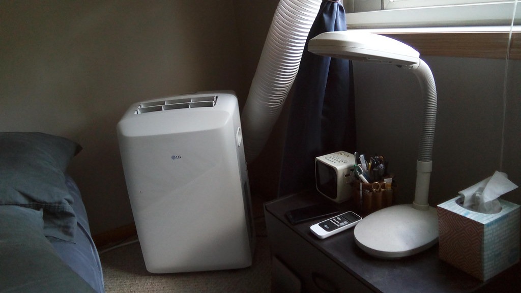White portable air conditioner on a bedroom carpet with exhaust hose running toward a curtained window